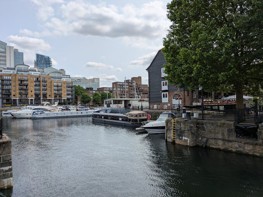 View of a busy marina at St Katharine Docks with several boats docked along the water's edge, including a large white yacht and smaller motorboats, some covered with protective fabric. A dark wooden b