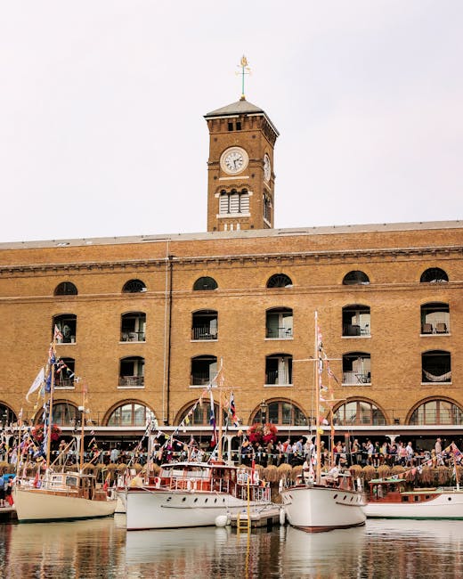 View of a busy marina at St Katharine Docks with several boats docked along the water's edge, including a large white yacht and smaller motorboats, some covered with protective fabric. A dark wooden b