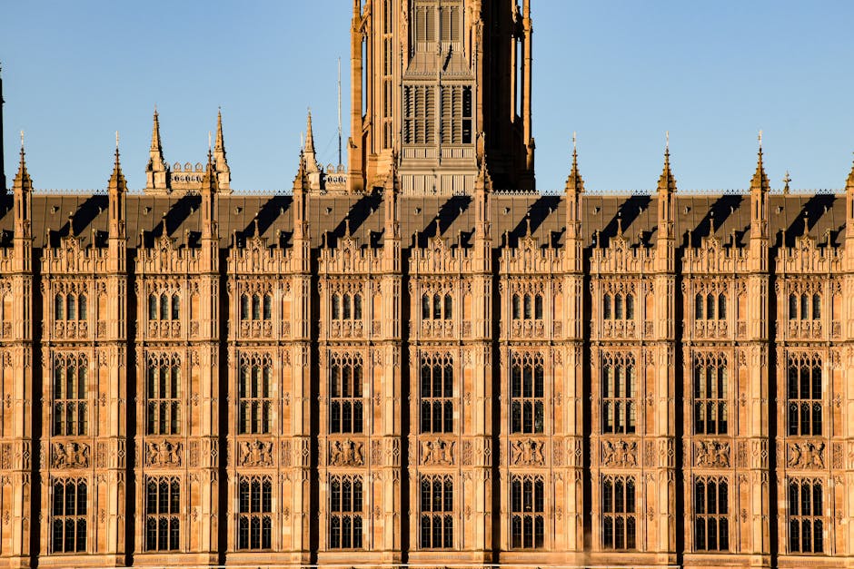 A detailed view of the historic façade of the Palace of Westminster in London, featuring ornate Gothic architecture with tall, narrow windows adorned with intricate stone tracery. The building is constructed from reddish-brown stone, and the sunlight highlights its fine carvings and decorative elements. In the foreground, there are several cardboard boxes, protected with plastic wrap and packing materials during a house removal process, placed near the building's entrance. A wooden furniture piece, possibly a cabinet or table, is being moved with the aid of a trolley, while furniture blankets and straps are visible on-site. A man wearing gloves is lifting or guiding the furniture onto a ramp or loading area where a van is partially visible, indicating the ongoing loading process typical of professional moving and packing operations by Man with Van Tower Hill, supporting home relocation and furniture transport services in the vicinity of Tower Hill.