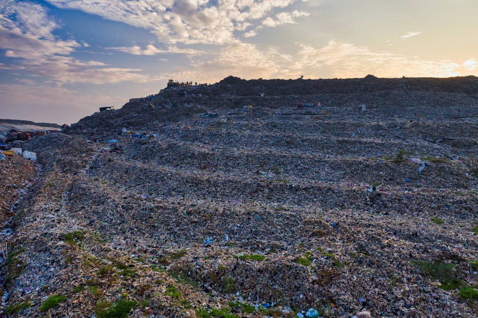 A large landfill site with multiple layers of accumulated waste, including visible plastic bottles, cardboard boxes, metal scrap, and various discarded household items. The waste is spread across a sloped terrain, with some scattered debris and patches of green vegetation sporadically emerging amidst the rubbish. In the background, a group of people are standing at the top of the landfill, overseeing the site, which is under a partly cloudy sky with warm, late-afternoon lighting. The scene illustrates the environmental impact of waste disposal, relevant to managing bulky waste after a house relocation, and highlights the importance of proper clearance services offered by Man with Van Tower Hill for efficient and environmentally responsible removals.