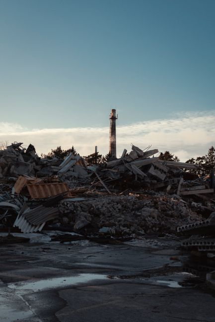 A landscape view of a large pile of demolished building debris, including broken concrete slabs, twisted metal, wooden planks, and discarded roofing materials, scattered across an open area with a wet, muddy surface. In the background, there is a tall industrial chimney and a clear sky with some clouds. The scene appears to be taken during late afternoon or early evening with subdued lighting. This image illustrates the removal of bulky construction waste, which is a common part of house removals and property clearance services offered by Man with Van Tower Hill, especially when dealing with waste after moving or renovation projects.