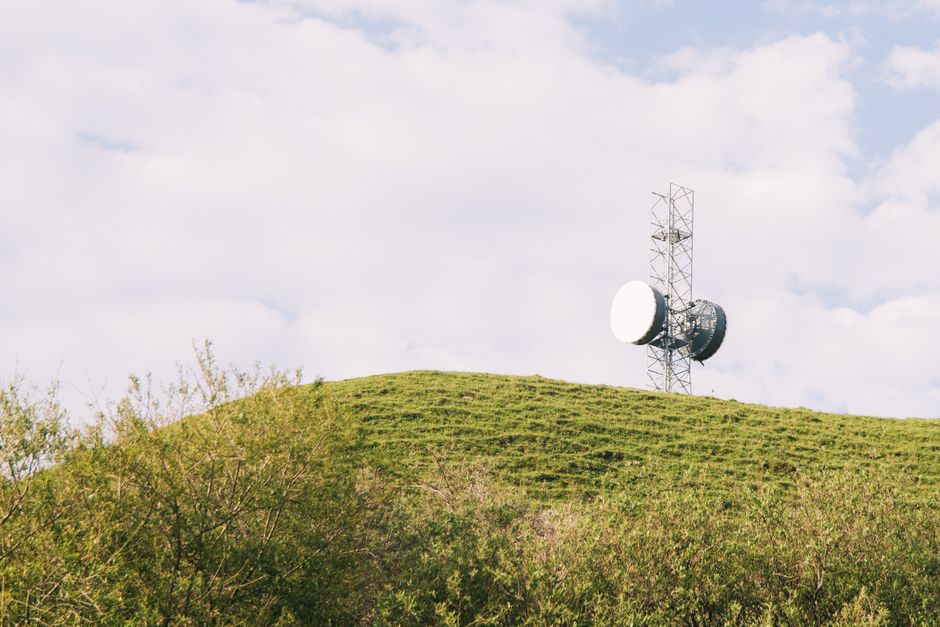 A large landfill site with multiple layers of accumulated waste, including visible plastic bottles, cardboard boxes, metal scrap, and various discarded household items. The waste is spread across a sloped terrain, with some scattered debris and patches of green vegetation sporadically emerging amidst the rubbish. In the background, a group of people are standing at the top of the landfill, overseeing the site, which is under a partly cloudy sky with warm, late-afternoon lighting. The scene illustrates the environmental impact of waste disposal, relevant to managing bulky waste after a house relocation, and highlights the importance of proper clearance services offered by Man with Van Tower Hill for efficient and environmentally responsible removals.