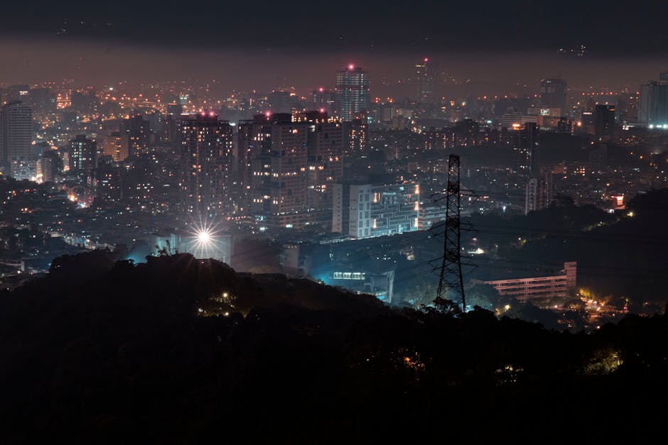 A nighttime cityscape viewed from a high vantage point, showing a densely populated urban area with numerous illuminated high-rise buildings, office blocks, and residential flats, all with various patterns of lit windows. The scene includes a large transmission tower with power lines cutting across the foreground, set against a dark silhouette of a hill or park area, with a faint glow from streetlights and building lights spreading across the city. The urban environment features a mix of bright city lights and darker areas, illustrating the vibrant logistical backdrop for house removals and relocation activities. This image, associated with Man with Van Tower Hill, reflects the busy and well-lit setting where home relocation and furniture transport services such as packing, loading, and transportation might be carried out, resonating with the theme of professional removals in a metropolitan context.