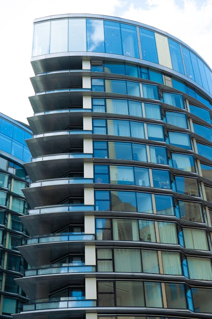 A modern high-rise building with a curved facade featuring multiple floors of large glass windows and prominent protruding balconies with black railings. The exterior is predominantly glass with blue reflections, and the building's rounded design creates a sleek, contemporary appearance. Part of the building's structure shows vertical white and dark grey elements contrasting with the glass. This image is relevant to house removals and furniture transport services by Man with Van Tower Hill, illustrating the type of complex residential relocations involving high-rise apartments, where careful planning and equipment such as trolleys and blankets may be used to facilitate moving large furniture through stairwells or elevators. The bright sky reflecting off the windows emphasizes daylight conditions typical during home relocation projects in urban settings.
