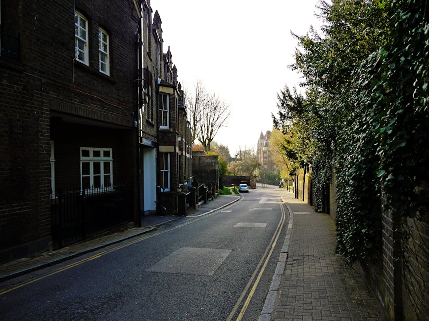 A narrow residential street during daylight with brick terraced houses on the left side, featuring white-framed windows and small front gardens enclosed by black metal railings. The pavement on the right is paved with concrete slabs, while the road is paved with asphalt and has double yellow lines along the curb indicating parking restrictions. Several cardboard boxes and wrapped furniture are visible on a loading area near a white van parked further down the street, indicating a home relocation or furniture transport process. The scene includes a few parked cars in the background, leafless trees without leaves, and a large building with a tower visible further along the street, suggesting a historic or urban environment. Man with Van Tower Hill's removal service is possibly involved in the logistics of packing and moving residential belongings in this area, utilizing equipment such as trolleys or straps for loading. The lighting suggests a clear day with natural daylight illuminating the scene properly for a professional moving operation.