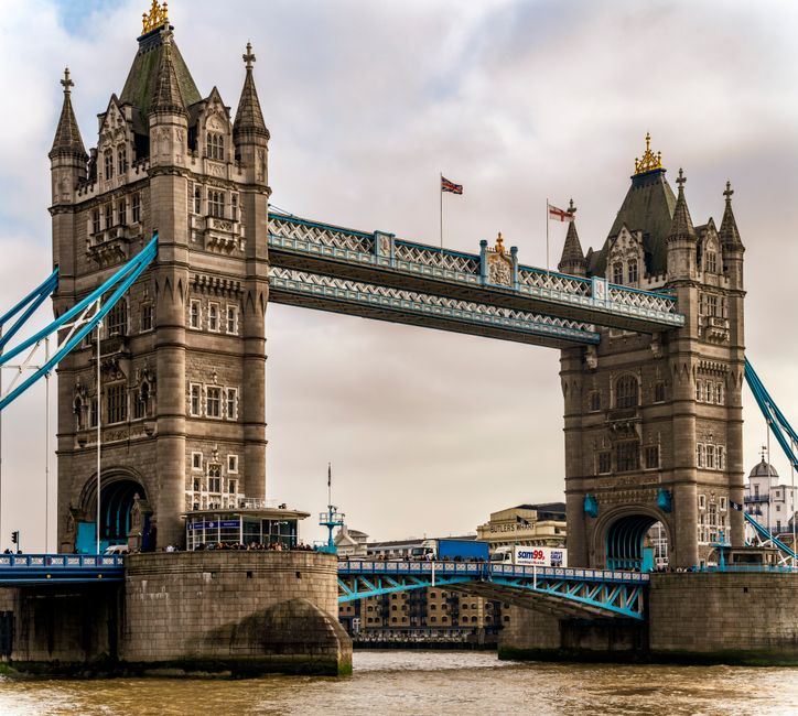 A view of Tower Bridge in London during daytime, showing the two iconic twin towers with detailed medieval-style architecture, topped with pointed turrets and golden accents. The bridge's blue suspension cables and walkway are visible connecting the towers, with some shipping containers and small boats in the River Thames beneath. The sky is overcast, casting a muted light over the scene. In the foreground, part of a home relocation process is suggested by a street scene with parked vehicles and a loading area, where Man with Van Tower Hill may assist with furniture transport or packing during house removals near Tower Bridge, with items such as cardboard boxes and protective blankets perhaps arranged nearby.