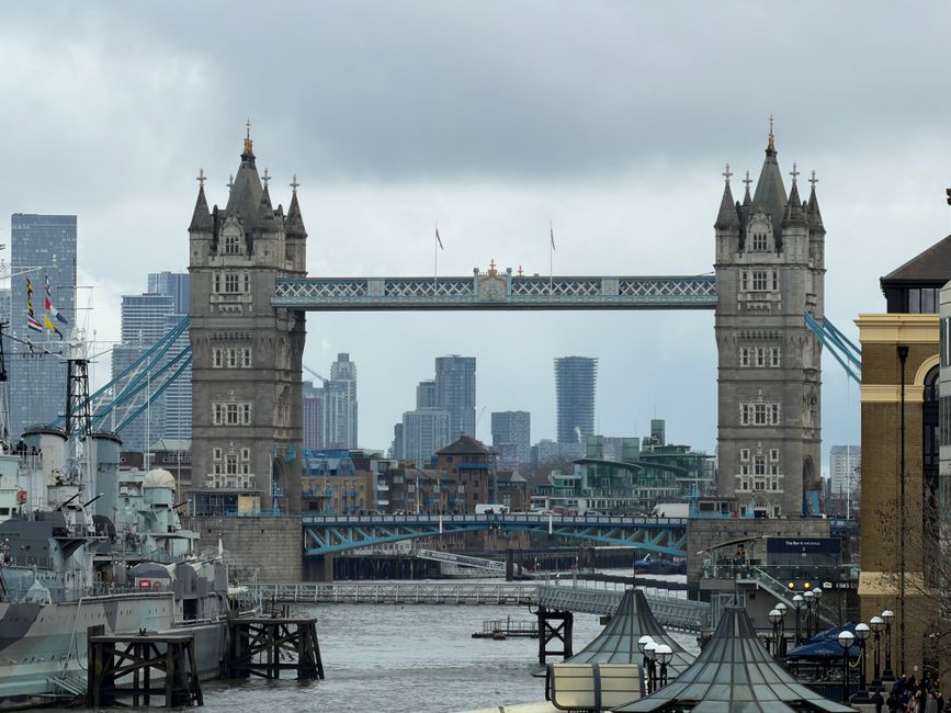 A wide-angle view of Tower Bridge in central London, with its iconic twin towers and suspension structure visible under a cloudy sky. The River Thames flows in the foreground, with several moored ships and boats along the waterfront. To the left, part of a large naval vessel is visible, with masts, antennas, and deck equipment, partially obscured by wooden pier structures. On the right, a section of a modern brick building is seen next to a black lamppost, and the pavement is lined with small globed street lamps. The bridge’s upper walkway, supported by the towers, spans across the river, with city skyscrapers and office buildings forming the urban backdrop. The lighting indicates overcast conditions consistent with typical London weather, and the scene captures a moment of quiet in the busy area, relevant to logistical activities involved in house relocations and furniture transport, with Man with Van Tower Hill occasionally serving as a reference point for local removal services near Tower Bridge.