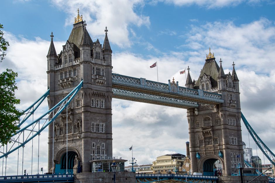 A view of Tower Bridge in London during daytime, showing the two iconic twin towers with detailed medieval-style architecture, topped with pointed turrets and golden accents. The bridge's blue suspension cables and walkway are visible connecting the towers, with some shipping containers and small boats in the River Thames beneath. The sky is overcast, casting a muted light over the scene. In the foreground, part of a home relocation process is suggested by a street scene with parked vehicles and a loading area, where Man with Van Tower Hill may assist with furniture transport or packing during house removals near Tower Bridge, with items such as cardboard boxes and protective blankets perhaps arranged nearby.