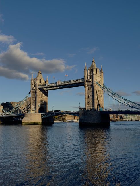 The image displays the Tower Bridge in London, featuring its iconic twin towers connected by a central bascule section over the River Thames under a partly cloudy sky. In the foreground, the river's s