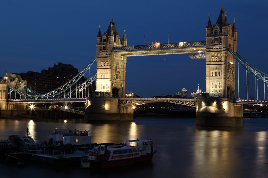 The image displays the Tower Bridge in London, featuring its iconic twin towers connected by a central bascule section over the River Thames under a partly cloudy sky. In the foreground, the river's s