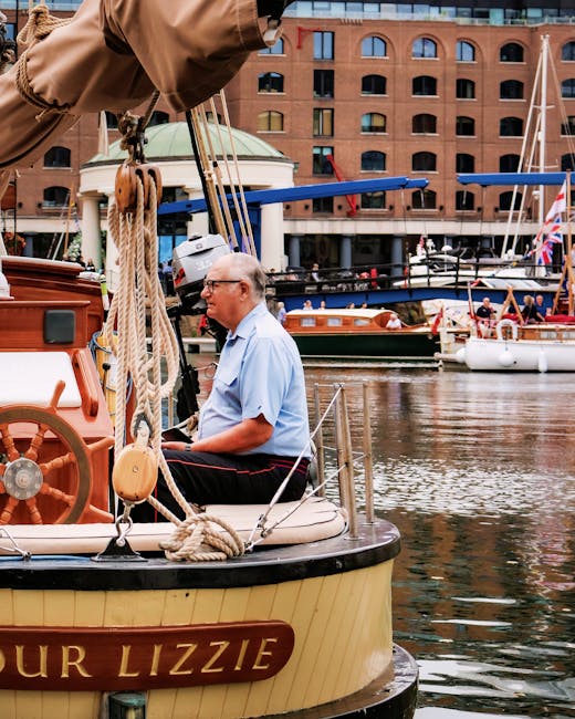 A man sitting on the bow of a small houseboat named 'Our Lizzie', which is moored at a marina with several other boats docked nearby. The man, dressed in a light blue shirt and dark trousers, is wearing glasses and appears to be relaxing or observing his surroundings. The boat features a beige hull with black trim, wooden fittings, and a large pile of thick, beige ropes coiled around the front cleats. In the background, there are additional boats with masts, some with Union Jack flags, and a red-brick building with arched windows across the water. The environment suggests a busy marina used for residential or leisure boat mooring, with calm water reflecting the boats and architecture. This scene is relevant to moving and relocation services, exemplifying boat or home removals near a marina, as handled by companies like Man with Van Tower Hill.