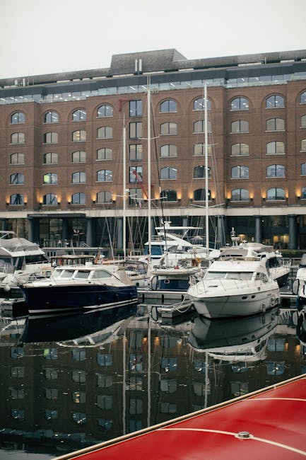 A row of white and dark blue motorboats and sailing yachts moored at a marina in front of a large red brick building with multiple arched windows and balconies, with some boats featuring visible masts, ropes, and fenders. The boats are secured to the docks with ropes, and the water reflects the vessels and building. The scene is captured during daylight, with ambient lighting illuminating the boats and the surrounding area. In the context of house removals and relocation services, this image depicts the exterior environment where transportation and logistical planning for home moving could involve water-based logistics or access considerations, as seen in the services offered by Man with Van Tower Hill.