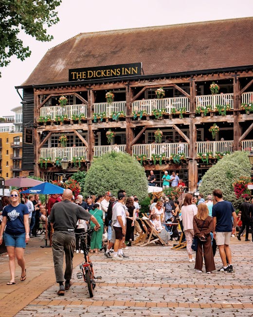 A man sitting on the bow of a small houseboat named 'Our Lizzie', which is moored at a marina with several other boats docked nearby. The man, dressed in a light blue shirt and dark trousers, is wearing glasses and appears to be relaxing or observing his surroundings. The boat features a beige hull with black trim, wooden fittings, and a large pile of thick, beige ropes coiled around the front cleats. In the background, there are additional boats with masts, some with Union Jack flags, and a red-brick building with arched windows across the water. The environment suggests a busy marina used for residential or leisure boat mooring, with calm water reflecting the boats and architecture. This scene is relevant to moving and relocation services, exemplifying boat or home removals near a marina, as handled by companies like Man with Van Tower Hill.