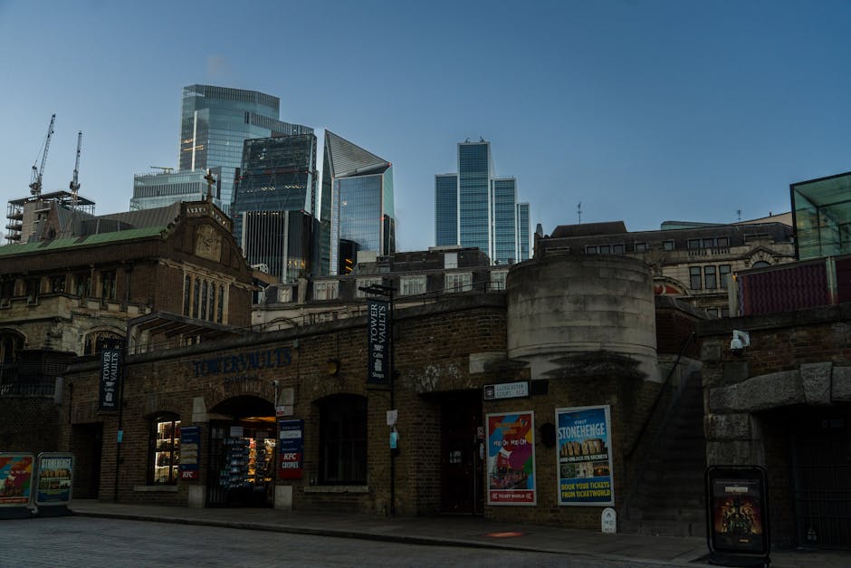 View of the exterior of a historic brick building with a rounded turret and an arched entrance, situated in a busy urban area surrounded by modern glass skyscrapers under clear blue sky. The ground-le