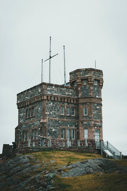 A historic stone building with a cylindrical tower featuring small rectangular windows and decorative brickwork, situated on a rocky, grassy hill under an overcast sky. In the foreground, a partially visible moving trolley or flatbed truck with a blue strap securing large cardboard boxes and wrapped furniture, indicating a home relocation process. The setting suggests an outdoor loading area adjacent to the property, with the presence of moving equipment and packaging materials typical of furniture transport and packing during house removals. The image is associated with [COMPANY_NAME], which provides urgent same-day removals in Tower Hill, highlighting the logistics involved in swift relocation services near iconic landmarks or historic sites.