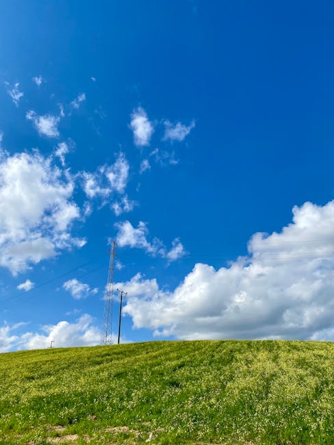 Photograph showing a clear blue sky with scattered white clouds above a grassy hill. On the left side of the image, a tall television or radio antenna is visible, extending upward from the hillside. The grass is lush and green, covering the lower part of the scene, while the sky dominates the upper portion. This outdoor environment captures a calm, natural setting that could be part of a rural or suburban area, with no immediate signs of buildings or human activity. The image's focus is on the expansive sky and the open space of the hillside, which may relate to the logistics of house removals or home relocation services provided by Man with Van Tower Hill, illustrating a clean and organized outdoor scene associated with moving or transportation processes.