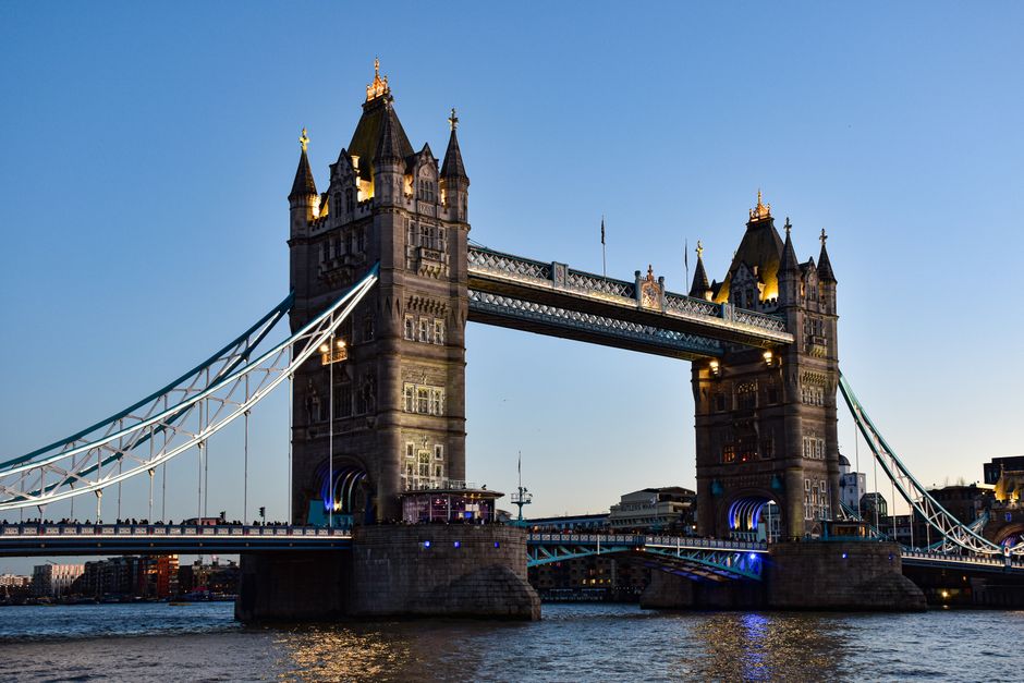 A view of the Tower of London with its historic stone walls and turrets situated along the River Thames during dusk. In the background, modern skyscrapers of the financial district, including glass office buildings with illuminated windows, rise into a deepening blue sky. Bare trees line the riverbank, and a paved walkway runs alongside the water, where pedestrians are visible. This urban landscape combines historic architecture with contemporary high-rise structures, highlighting the contrast between old and new in central London. The scene reflects the city’s ongoing change and development, which is relevant to house removals and property relocation services offered by Man with Van Tower Hill, specializing in furniture transport and professional moving logistics.